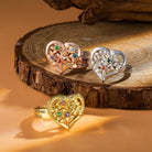 Three rings on a wooden surface: a gold one engraved with "Michael, Erica," a rose gold one with "Selena, Julian, Diane," and a silver one with "Calvin," all with heart and floral designs.