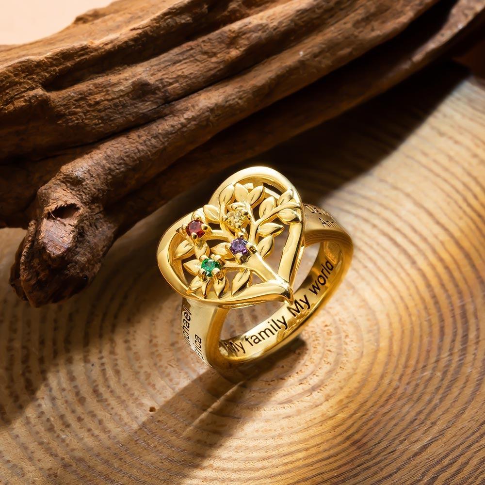 A gold ring with heart and floral design featuring colorful gems, engraved with "My family, My world," displayed on a wooden surface with a piece of wood in the background.