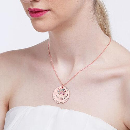 A woman wearing a strapless white dress and a rose gold family tree necklace with two disks, adorned with colorful gemstones and engraved names.