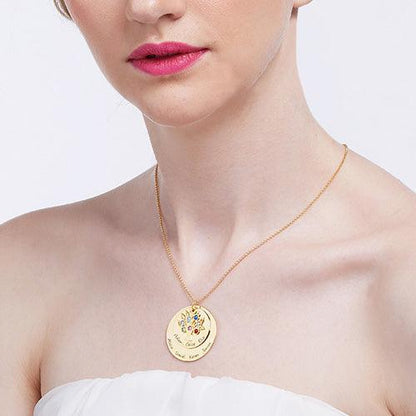 A woman wearing a strapless white dress and a gold family tree necklace with two disks, adorned with colorful gemstones and engraved names.