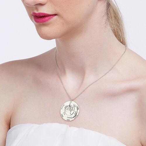 A woman wearing a silver circular pendant necklace with a tree design in the center, engraved with multiple names, against a white strapless top.