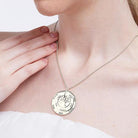 A woman touching a silver circular pendant necklace with a tree design in the center, engraved with multiple names, against a white strapless top.