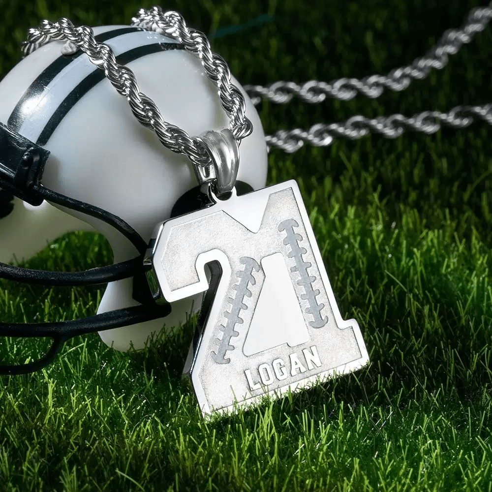 A silver number pendant with "21" and the name "Logan" engraved on it, hanging on a silver chain, placed on a miniature football helmet on green grass.