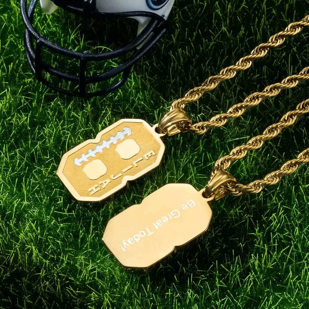 Two gold number pendants on chains, one showing "8" with the name "Elijah" and the other engraved with "Be Great Today!", placed on green grass near a miniature football helmet.