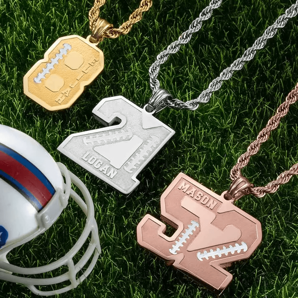 Three customized number pendants on chains: gold "8" for Elijah, silver "21" for Logan, and rose gold "52" for Mason, placed on green grass near a miniature football helmet.