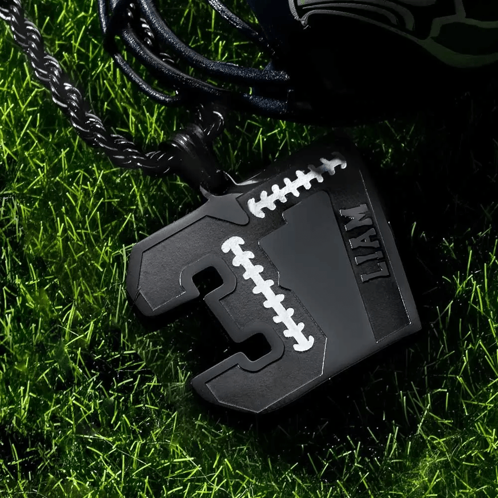 A black number pendant with "3" and the name "Liam" engraved on it, hanging on a black chain, placed on green grass near a miniature football helmet.