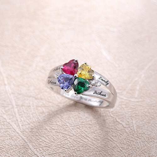 Silver ring with four heart-shaped multicolored gems on a textured beige background, engraved with names "Olivia," "Rebecca," "Sandy," "Joshua."