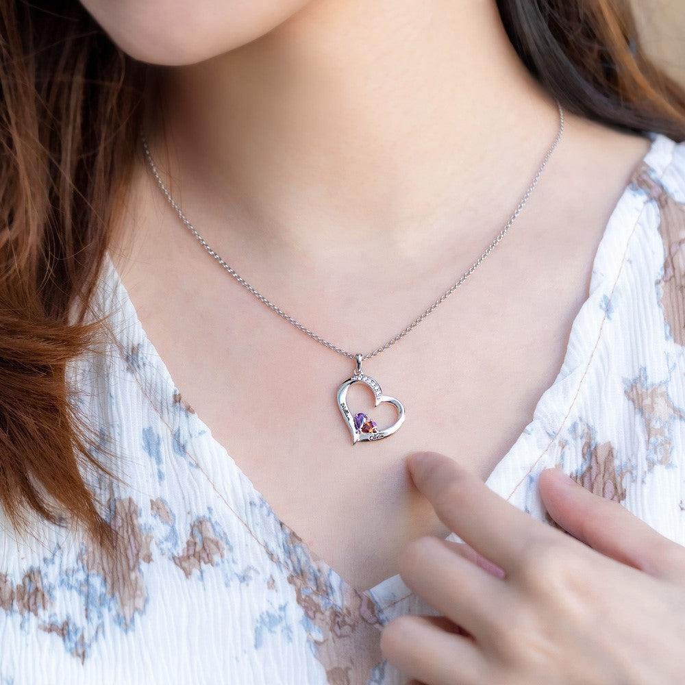 A woman wearing a heart-shaped pendant with colorful gemstones on a silver chain, touching the pendant, with a focus on the jewelry against her floral-patterned top.