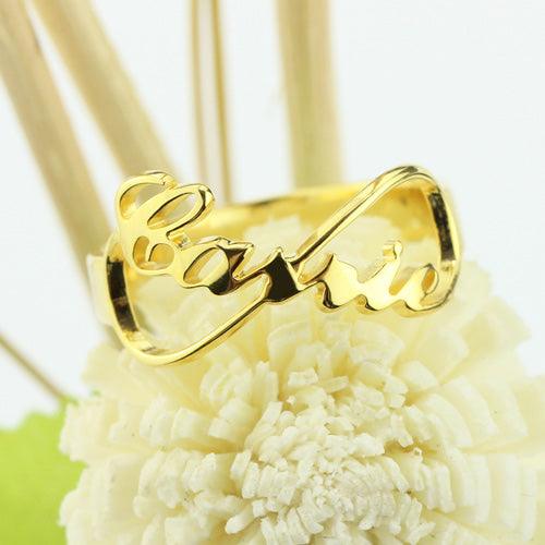 Gold ring with the word "Carrie" in cursive script, forming an infinity symbol design, placed on a decorative flower background.