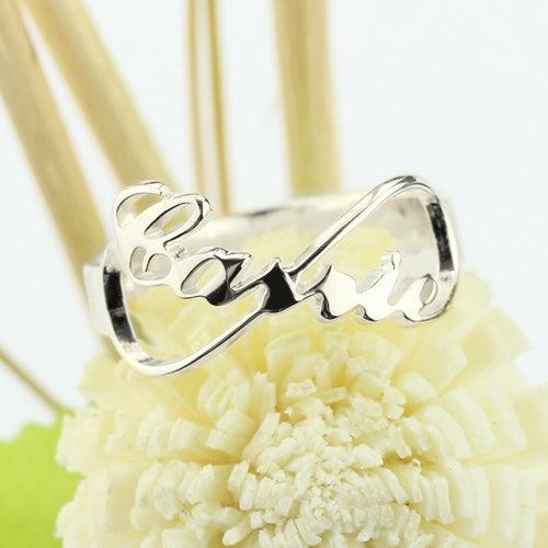 Silver ring with the word "Carrie" in cursive script, forming an infinity symbol design, placed on a decorative flower background.