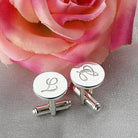 Silver cufflinks with engraved initial 'L' beside a pink rose on a textured grey surface.