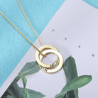 A gold necklace with two interlocking rings, engraved with the names "MELISSY" and "NITZAN", placed on a light blue and white background with leaf illustrations.