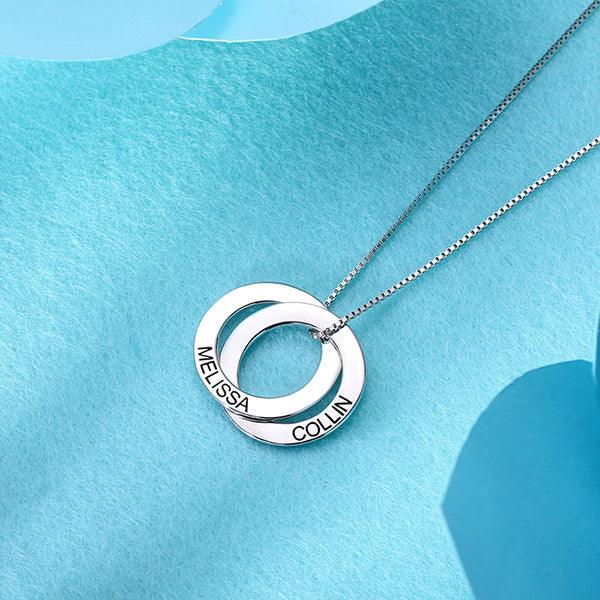 A silver necklace with two interlocking rings engraved with the names "MELISSA" and "COLLIN," placed on a textured light blue background.