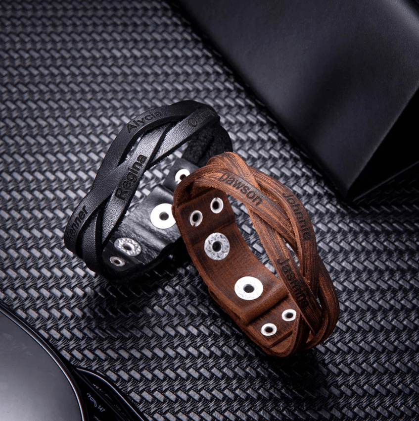 Two engraved leather bracelets, one black and one brown, with silver accents, on textured fabric.