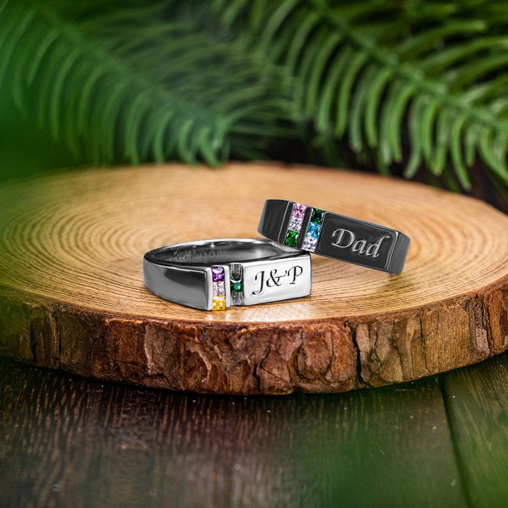 Two personalized rings with engraved names "Dad" and "J&P", featuring multicolored gemstones, displayed on a wooden surface with green foliage in the background.