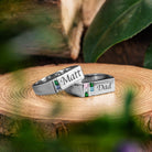 Two silver rings on a wooden surface, each engraved with the names "Matt" and "Dad," adorned with multicolored gemstones.