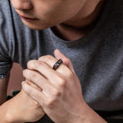 Man in a gray shirt clasping his hands, wearing a black ring engraved with "Dad" and accented with gemstones.