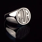 Elegant silver signet ring with 'MJA' initials monogram on a dark background with reflection.