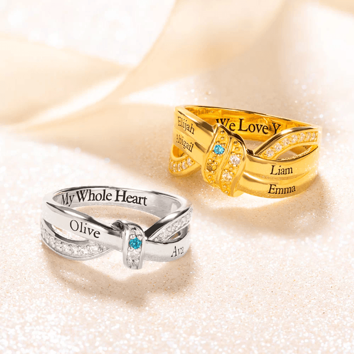 Two intertwined rings, one in silver and one in gold, engraved with names and adorned with small gemstones. The silver ring reads "My Whole Heart," and the gold ring reads "We Love You."