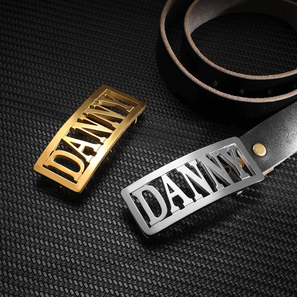 Gold and silver name belt buckles with 'DANNY' cut-out, beside a rolled-up brown leather belt on a textured black surface.