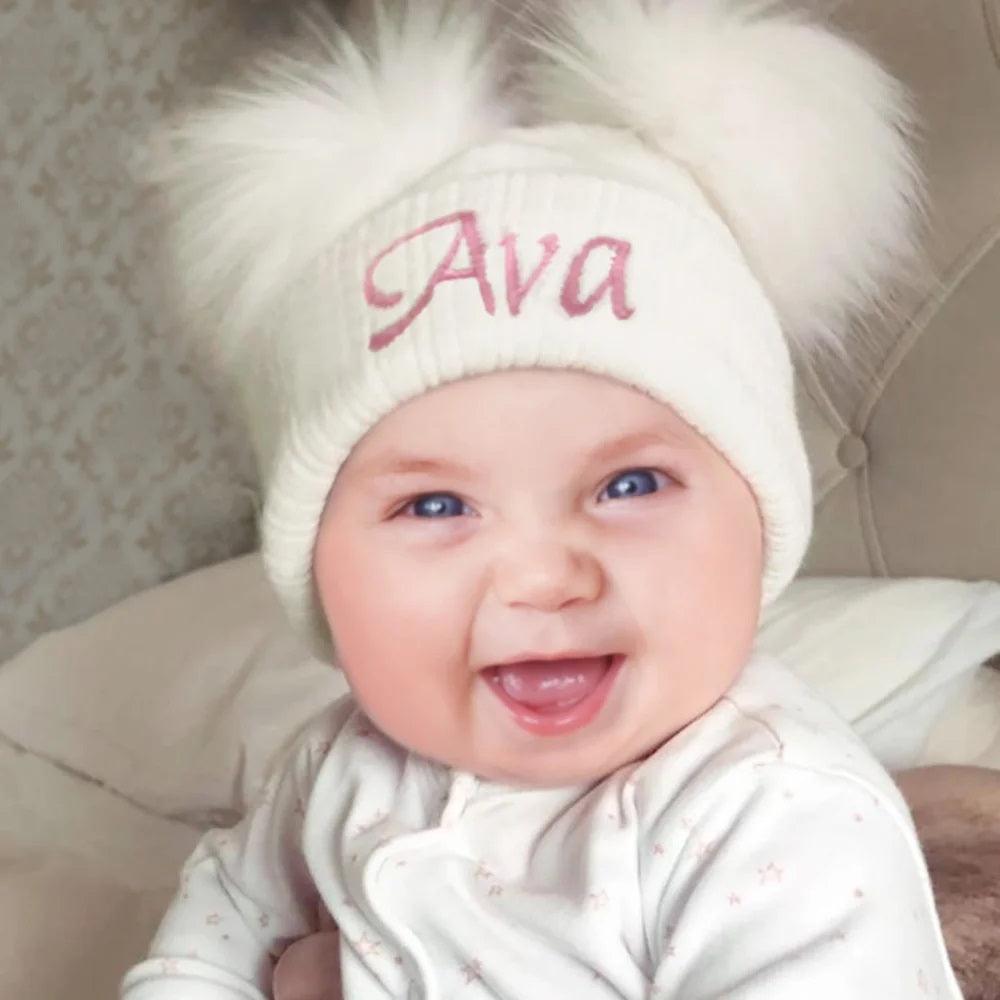 A joyful baby with bright blue eyes and a wide smile wears a white knit hat with fluffy pom poms, embroidered with the name "Ava" in pink.