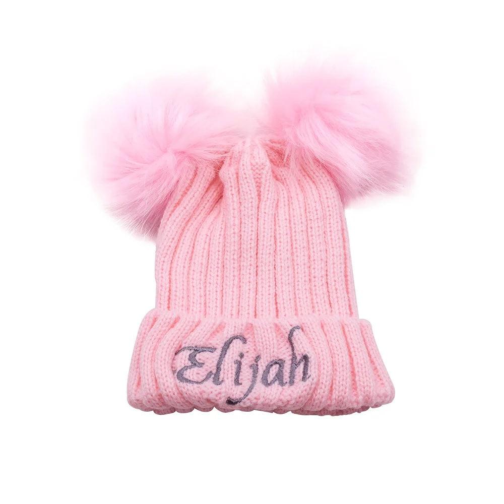 A pink knit baby hat with "Elijah" embroidered in cursive and two fluffy pink pom poms on top, isolated on a white background.