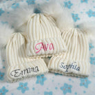 Three cream-colored knit baby hats with fluffy pom poms, each embroidered with a different name—Ava, Emma, Sophia—on a blue blanket with flower patterns.