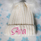 A cream-colored baby hat with a pink embroidered name "Ava" and two white pom poms, placed on a blue and white floral-patterned blanket.