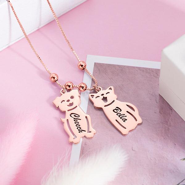 A rose gold necklace with two pendants shaped like a dog and a cat on a pink background. The dog pendant is engraved with "Cheech" and the cat pendant with "Bella."