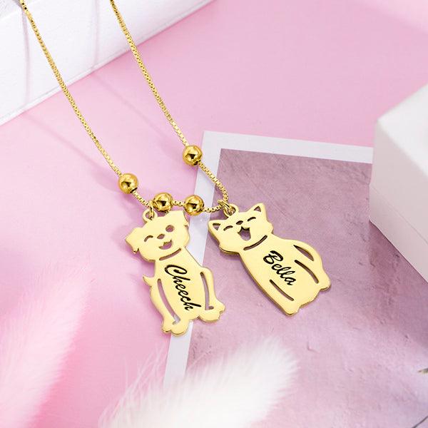 A gold necklace with two pendants shaped like a dog and a cat on a pink background. The dog pendant is engraved with "Cheech" and the cat pendant with "Bella."