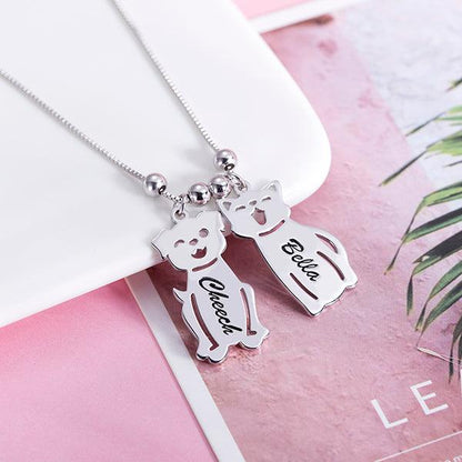 A silver necklace with two pendants shaped like a dog and a cat on a pink and white background. The dog pendant is engraved with "Cheech" and the cat pendant with "Bella."