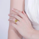 A close-up of a woman's hand wearing a gold ring with a tree of life design, highlighting the elegance of the jewelry against her light skin and white dress.