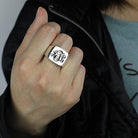 A person wearing a silver signet ring with a rectangular face, engraved with the initials "ATL" in an elegant, black cursive monogram style.