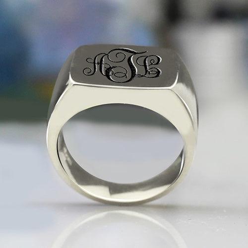 A silver signet ring with a rectangular face, engraved with the initials "ATL" in an elegant, black cursive monogram style, placed on a reflective surface.