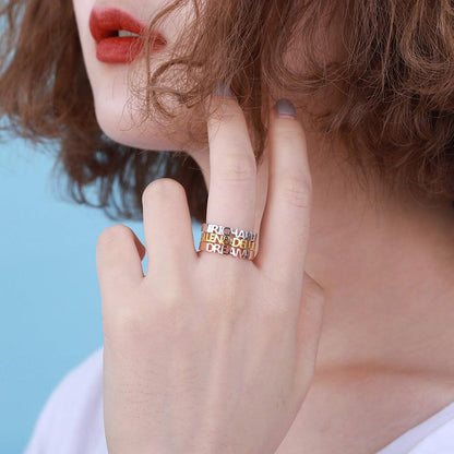 A close-up of a person with short, curly hair and red lipstick, wearing three rings in silver, gold, and rose gold with text on them, and holding their hand near their face.