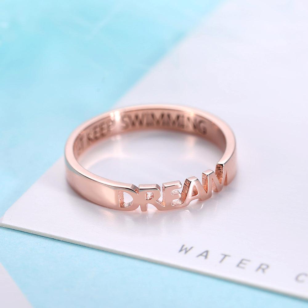 A rose gold ring with cut-out lettering that reads "DREAM." The inside of the ring is inscribed with the phrase "JUST KEEP SWIMMING."