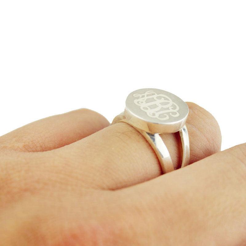 A silver ring with a semi-split shank band and an oval face engraved with intertwined initials "ABC" in cursive script, worn on a person's finger, giving it an elegant and distinctive appearance.