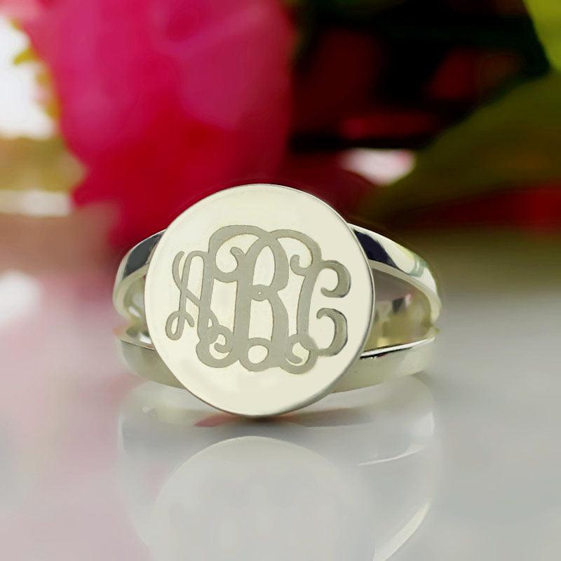 A silver ring with a semi-split shank band and an oval face engraved with the intertwined initials "ABC" in cursive script, displayed against a blurred floral background.