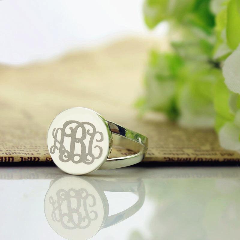 A silver ring with a semi-split shank band and an oval face engraved with the intertwined initials "ABC" in cursive script, placed on a reflective surface with a floral background.