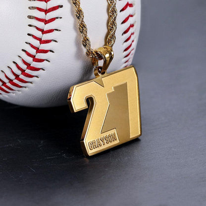 A gold pendant with the number 27 and the name "Grayson" engraved on it, hanging from a gold chain, placed against a white baseball with red stitching.