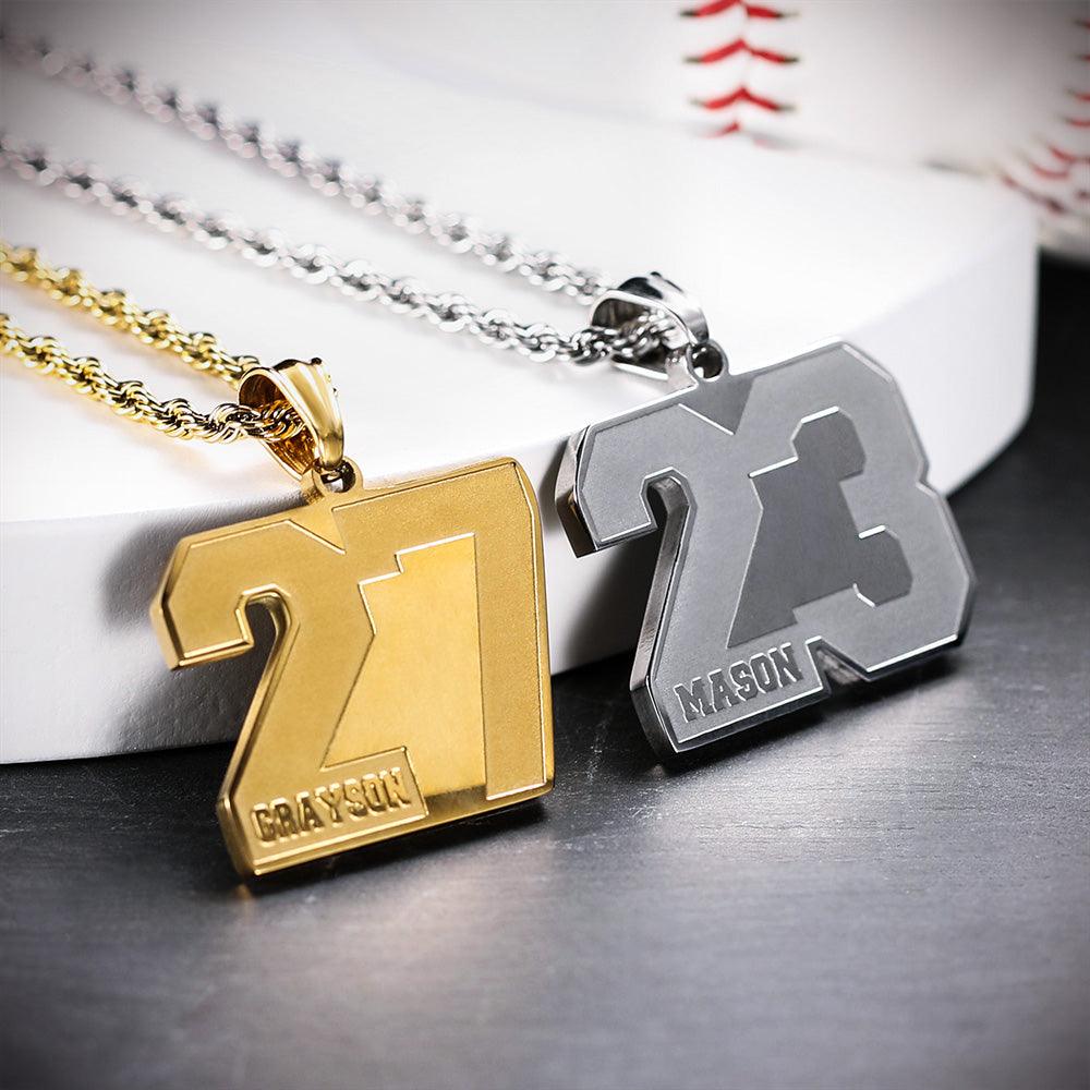 Two pendants, one gold with the number 27 and "Grayson," and one silver with the number 23 and "Mason," hanging from chains, placed near a baseball.