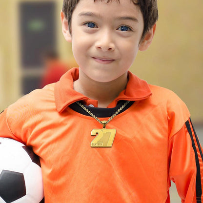 A young boy in an orange sports jersey holding a soccer ball, wearing a gold pendant with the number 27 and the name "Grayson" engraved on it.