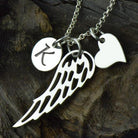 A silver necklace with three charms: a circular disc with the letter "K," an angel wing, and a heart, displayed on a dark, textured background.