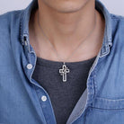 A person wearing a silver cross pendant with a hollow design and a ring inscribed with "LOVE." The pendant hangs from a silver chain over a gray shirt and denim jacket.