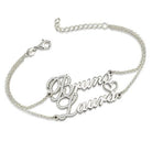 A  Silver bracelet with the names "Bruno" and "Laura" in cursive, featuring birthstones just before the letters B and L, connected by a delicate chain with small diamonds and a heart.