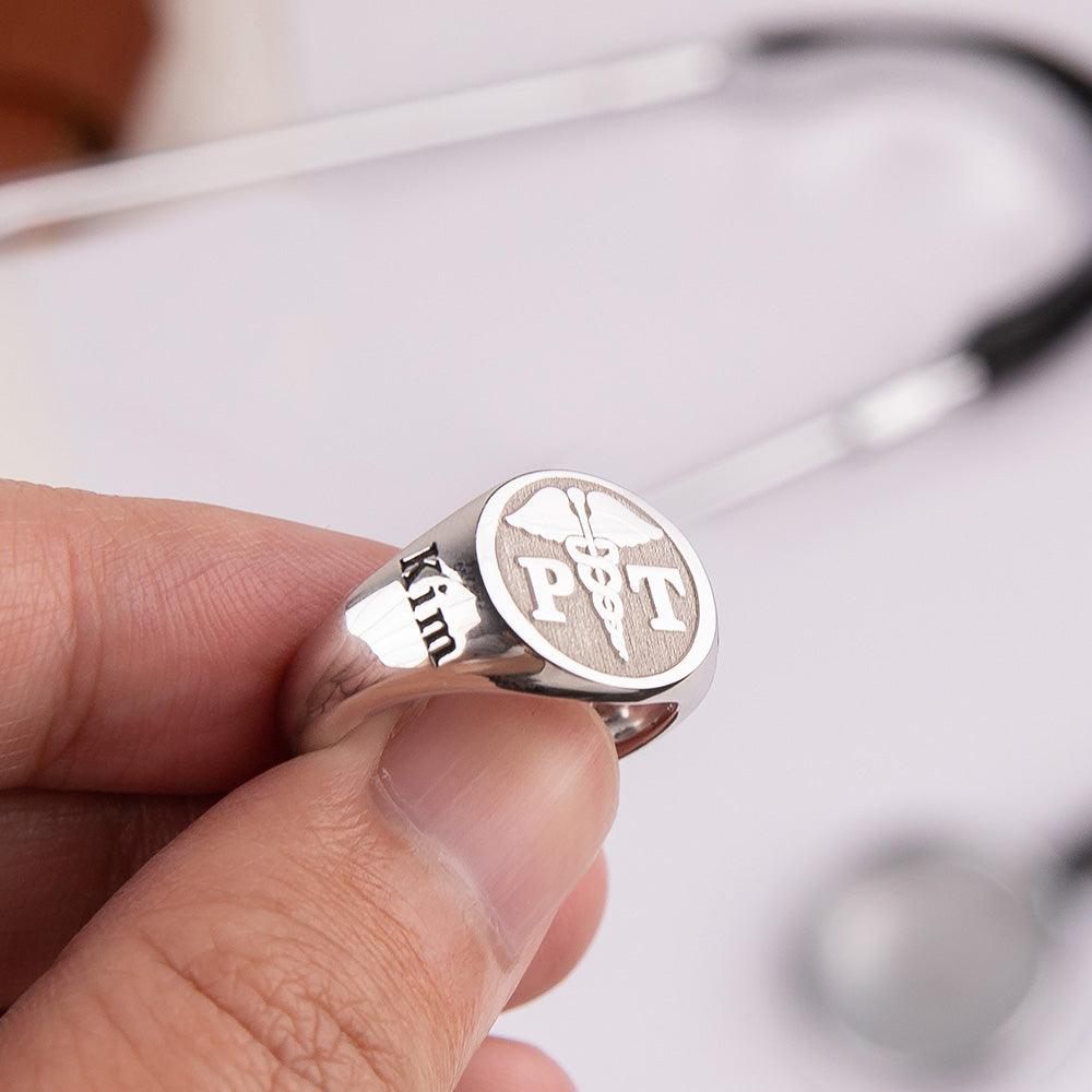 A close-up of a hand holding a silver ring engraved with a caduceus symbol and the letters "PT." The name "Kim" is also engraved on the ring.