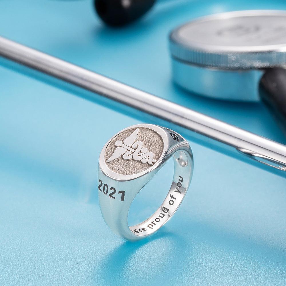 A silver ring with a caduceus symbol and the year "2021" engraved. The ring is inscribed with "We're proud of you" and displayed against a blue background with medical tools.