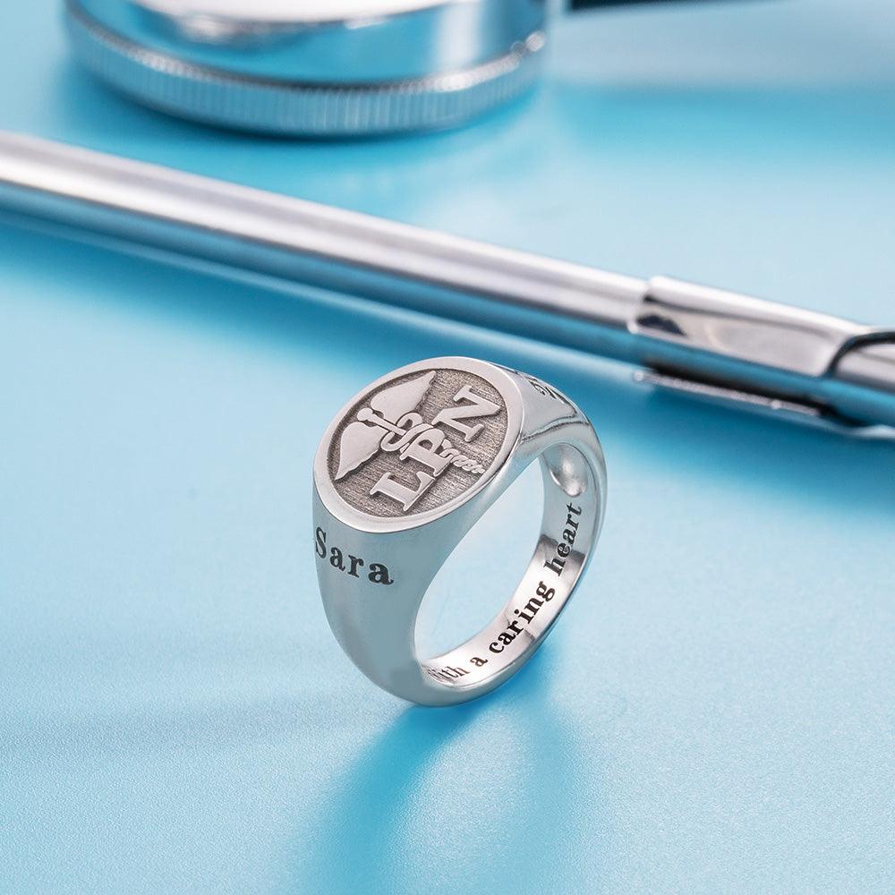 A silver ring with a caduceus symbol and the letters "RN" engraved. The ring also features the name "Sara" and the phrase "With a caring heart," displayed with medical tools on a blue background.