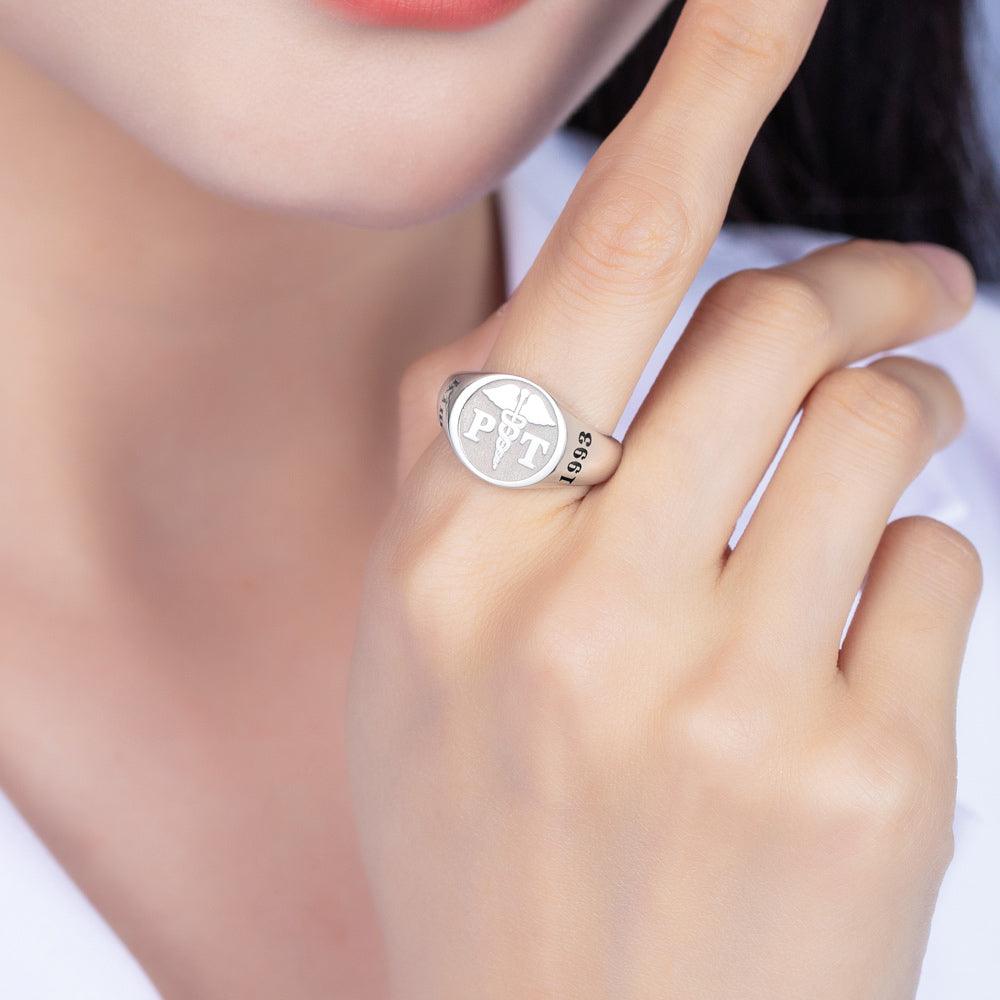 A person wearing a silver ring engraved with a caduceus symbol and the letters "PT." The ring also features the year "1983." The person is touching their face.