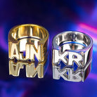 Two custom name rings, one in gold featuring 'AJN' and one in silver featuring 'KR,' displayed against a vibrant purple background with reflective surface.